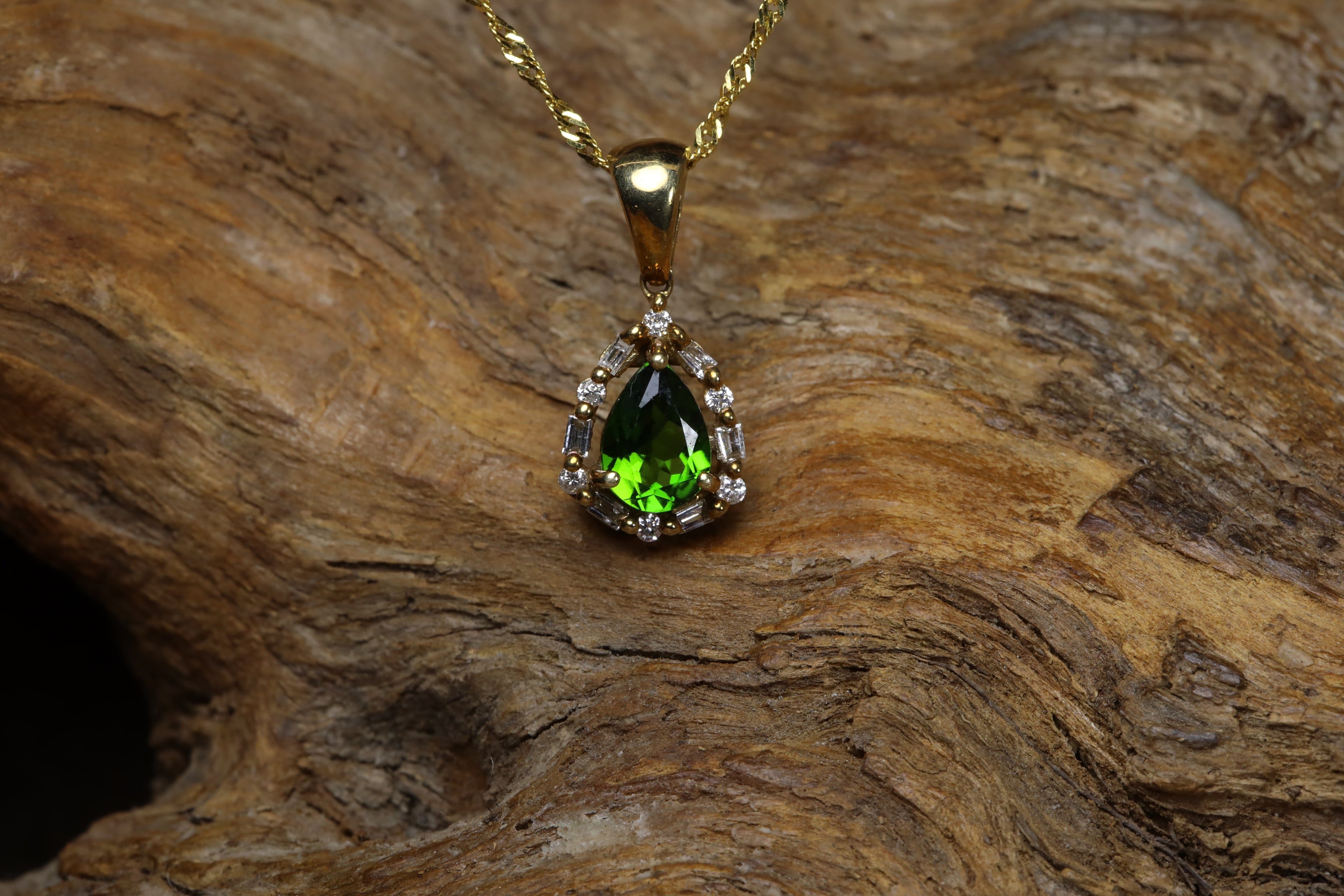 Vintage 9ct yellow gold pendant featuring a vivid green pear cut diopside stone surrounded by a halo of round and baguette diamonds, hanging on a gold chain against rustic wood