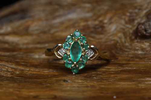 Vintage 9ct yellow gold ring featuring a marquise cut emerald centre stone surrounded by round emeralds in a diamond shape cluster setting with diamond shoulder accents, resting on rustic wood