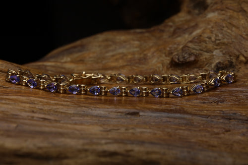 Vintage 9ct yellow gold tennis bracelet featuring pear cut violet-blue tanzanite stones set in claw settings with gold bar links, draped over rustic wood