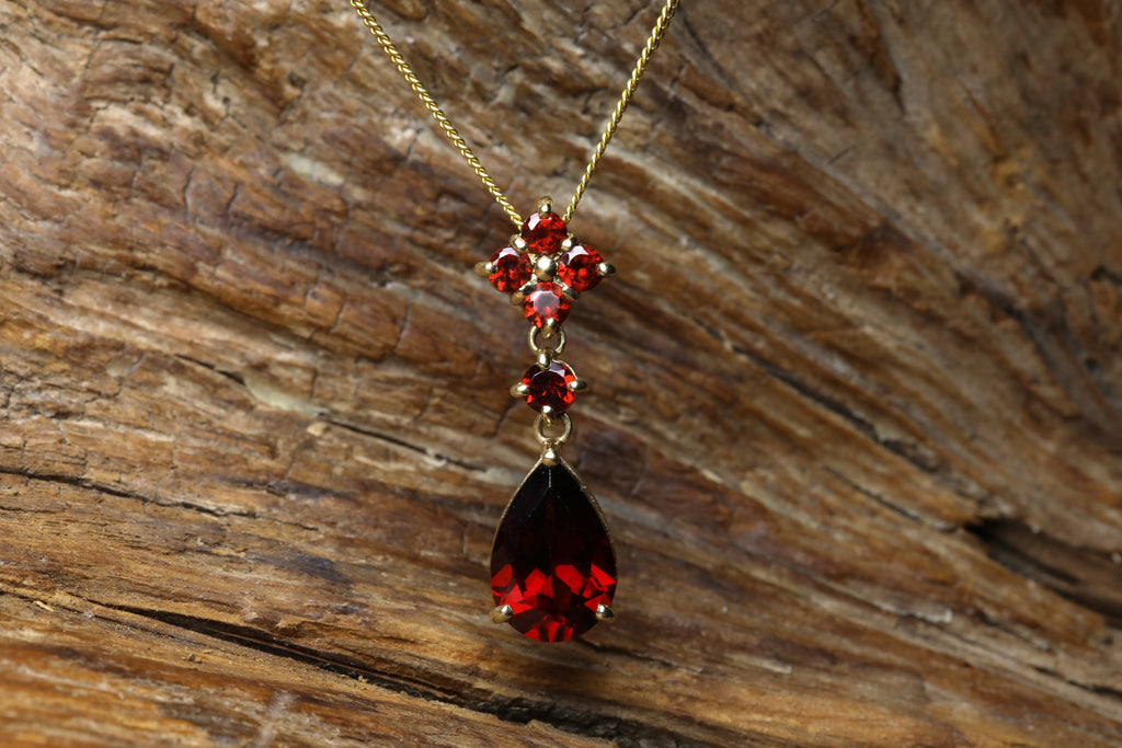 Vintage 9ct yellow gold garnet drop pendant featuring a pear cut deep red garnet with round garnet cluster top, hanging on a gold chain against a rustic wood background